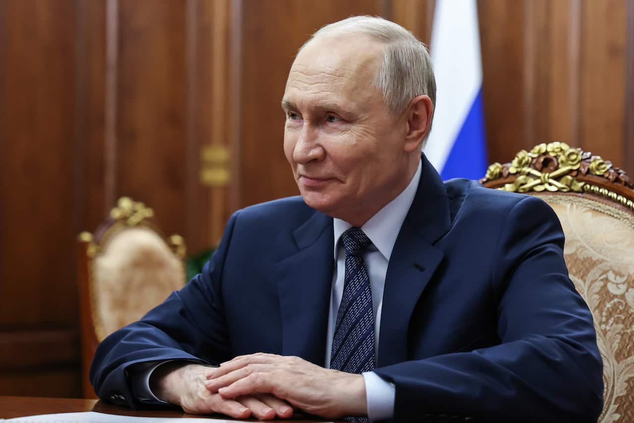 Man in a dark suit and tie sits at an ornate table in a wood-panelled room, with a Russian flag visible in the background.