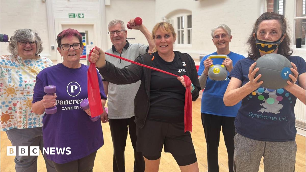 A group of middle-aged men and women holding gym equipment stand together in an exercise class.