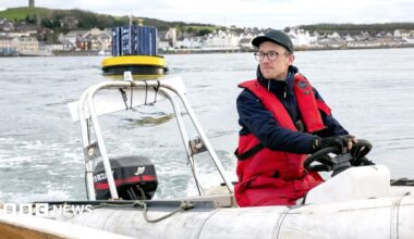 Man in a red flotation suit operates a small inflatable workboat in a harbour, with a floating marine device and waterfront buildings visible in the background.