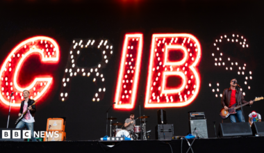The band The Cribs play on a big stage. Lights behind spell the band name.