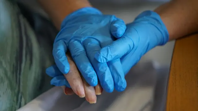 Someone wearing blue latex gloves holding the hands of someone in a hospital bed. On their hands and the edge of the bed are visible