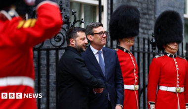 MI5 chief Sir Ken McCallum has short dark hair, dark glasses and is wearing a dark suit with a green tie.