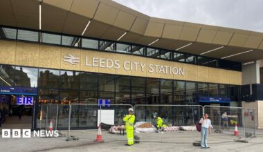 A railway station concourse. Workers in high vis mill about on the concourse and behind a temporary metal works barrier. A big sign in white lettering on a gold background indicates it's Leeds City Station. In the background we can see inside the station, with digital train times board visible, and people inside,