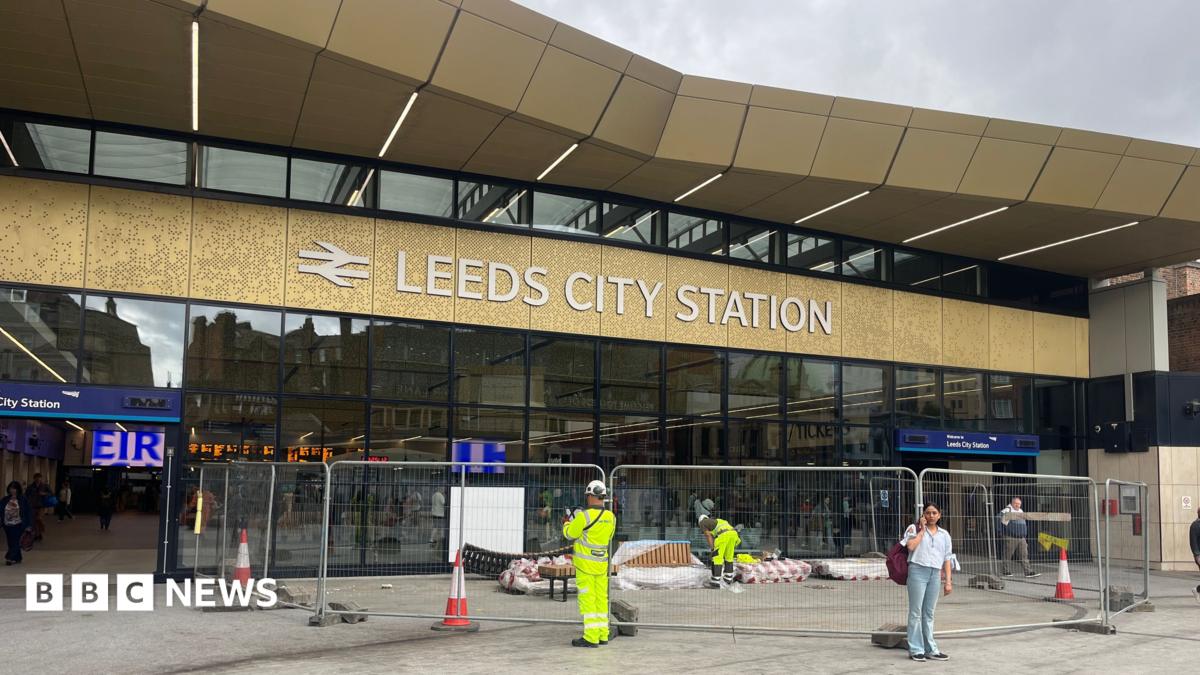 A railway station concourse. Workers in high vis mill about on the concourse and behind a temporary metal works barrier. A big sign in white lettering on a gold background indicates it's Leeds City Station. In the background we can see inside the station, with digital train times board visible, and people inside,