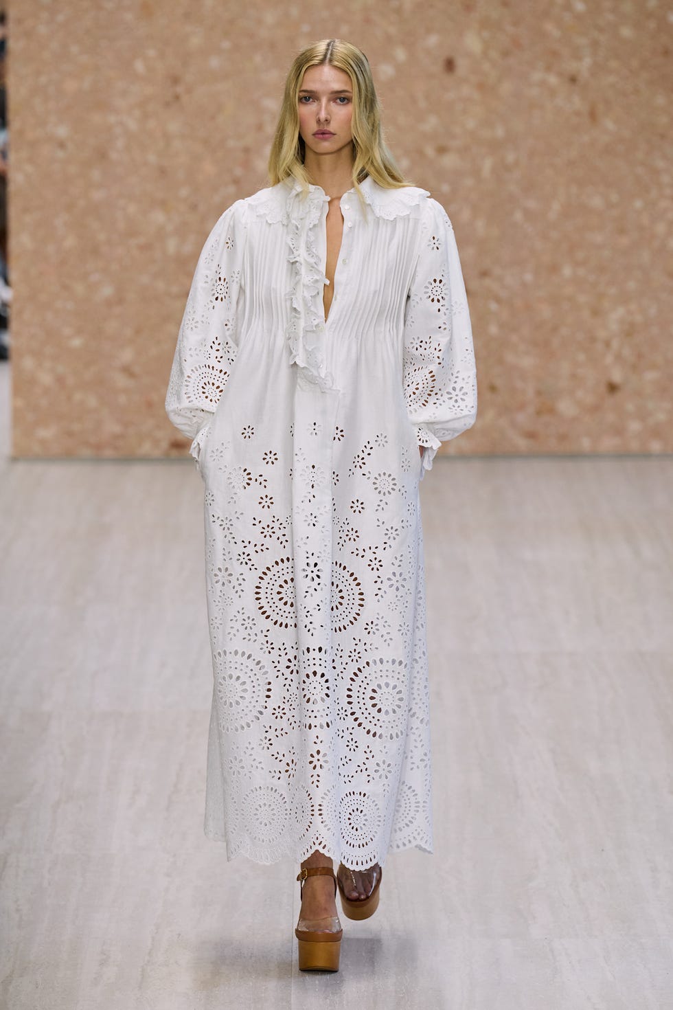 Model wearing a long white dress with intricate cut-out patterns and brown platform sandals on a runway.