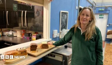 A woman is standing in a community hall, next to some cakes. She's wearing a green top and smiling.