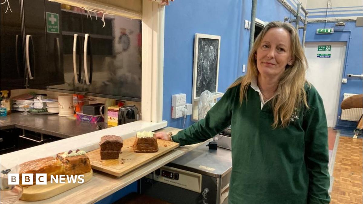 A woman is standing in a community hall, next to some cakes. She's wearing a green top and smiling.