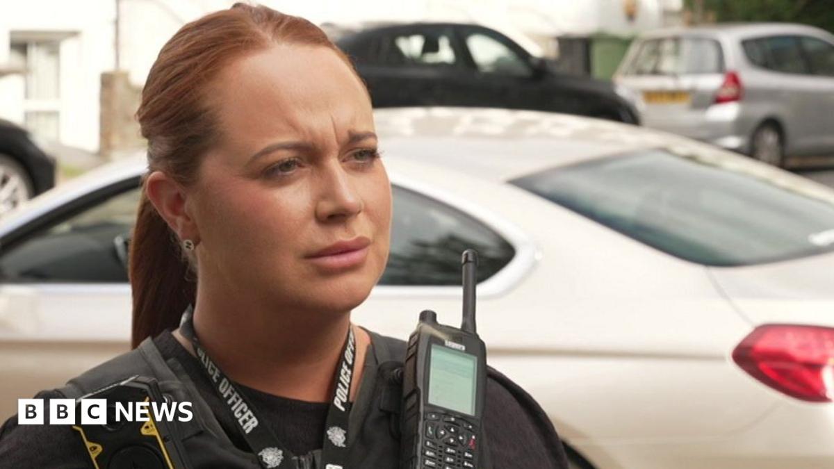PC Zoe Ticehurst in the street in front of rows of parked cars. She wears a protective vests and police radio
