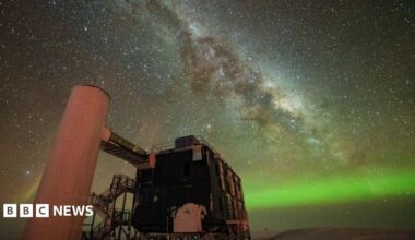 A starry sky the Milky Way is seen through the middle, and the Northern Lights in green along the horizon. In the foreground is an observatory with a black building and stairs liking to a white column.