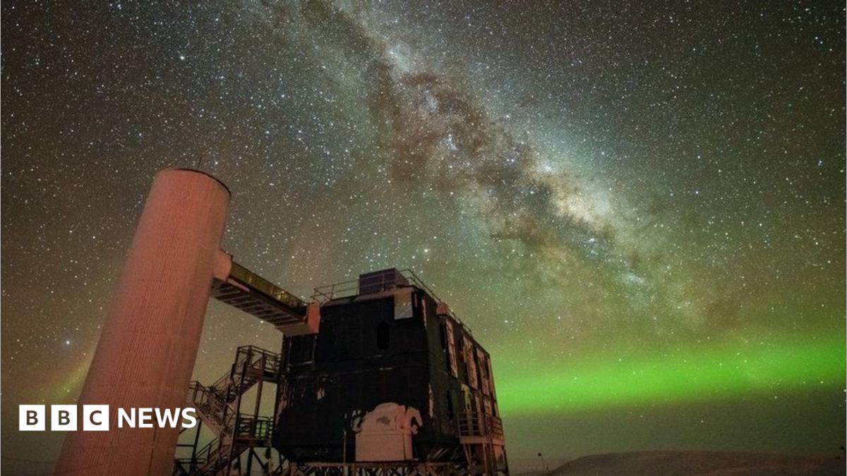 A starry sky the Milky Way is seen through the middle, and the Northern Lights in green along the horizon. In the foreground is an observatory with a black building and stairs liking to a white column.