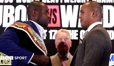 Daniel Dubois is wearing a blue jacket and has the WBO heavyweight title on his shoulder as he looks at Fabio Wardley, who is wearing a brown jacket. Frank Warren, in a tan jacket, stands between the pair, partly in shadow.