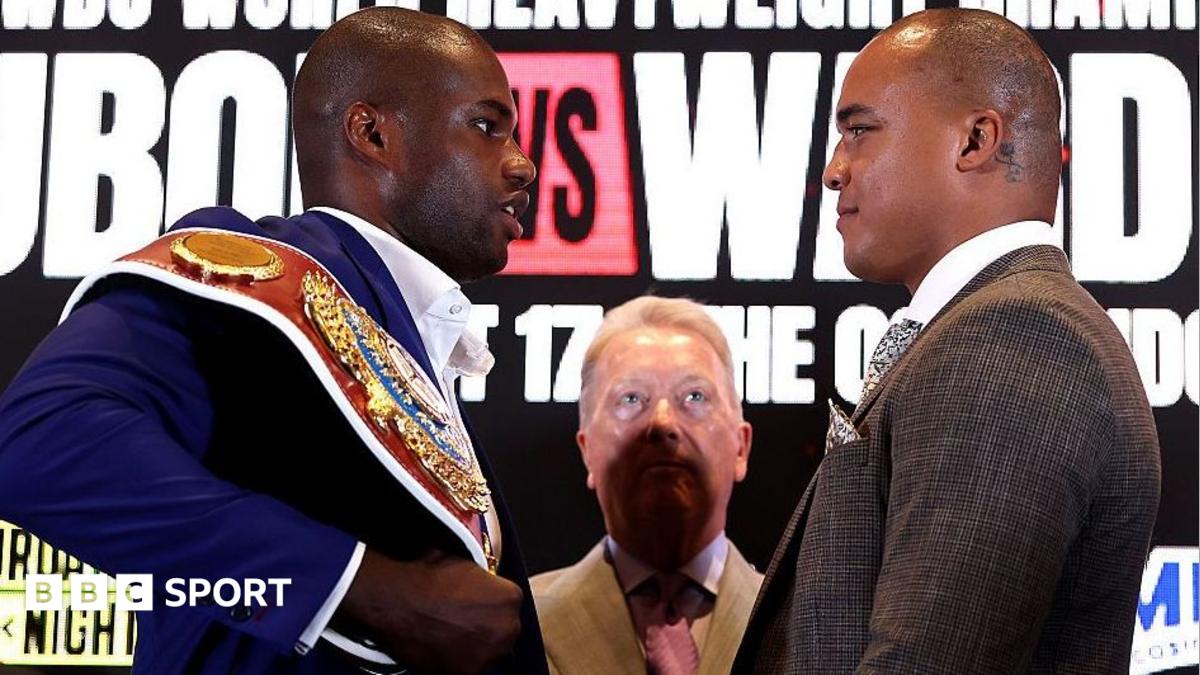 Daniel Dubois is wearing a blue jacket and has the WBO heavyweight title on his shoulder as he looks at Fabio Wardley, who is wearing a brown jacket. Frank Warren, in a tan jacket, stands between the pair, partly in shadow.