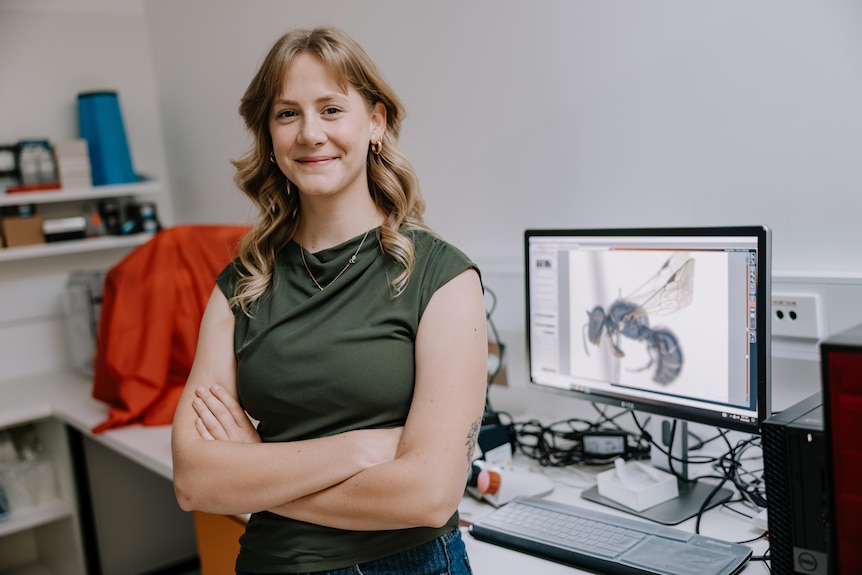 A smiling woman with golden-brown hair stands with her arms crossed in front of a computer in an office.