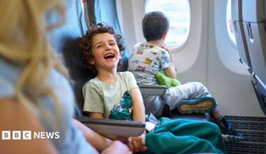 A child laughs while sitting on a plane next to a younger sibling on one side and parent on the other