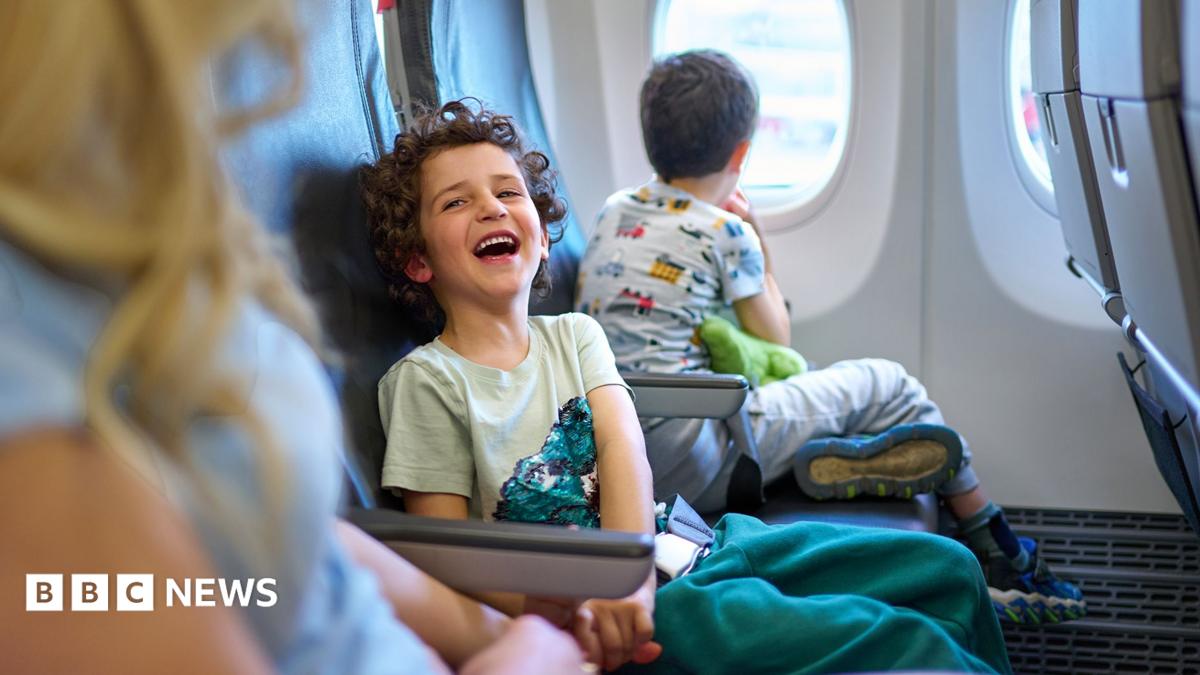 A child laughs while sitting on a plane next to a younger sibling on one side and parent on the other