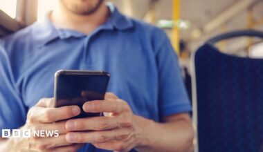 A man in a blue Tshirt holding a mobile phone and sitting on a bus
