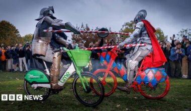 Two men dressed in medieval knight regalia ride lime bikes and hold red and silver striped poles as they attempt to knock each other off their bikes.