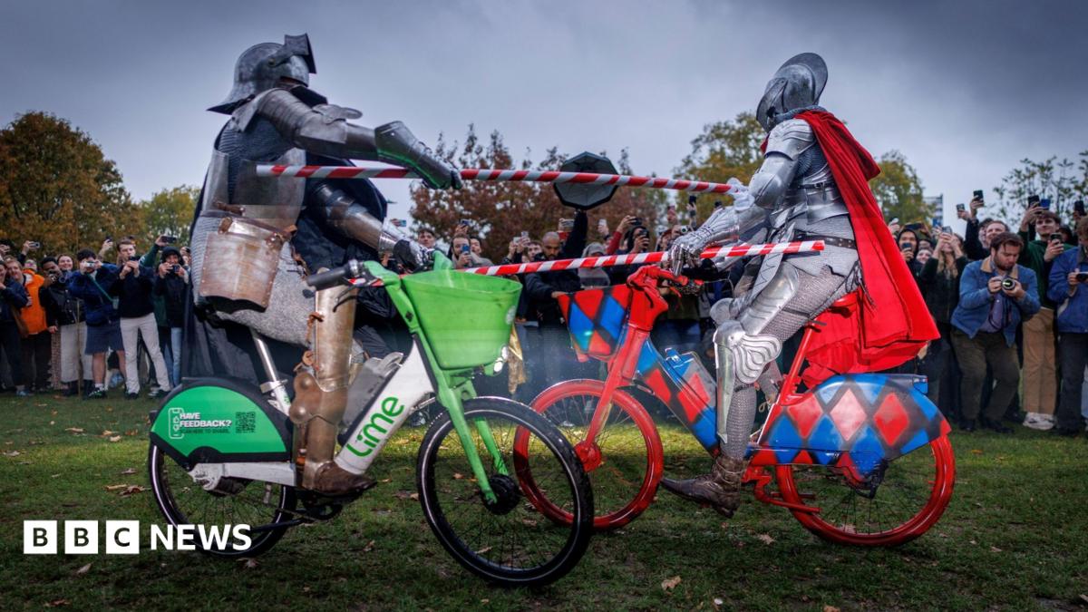 Two men dressed in medieval knight regalia ride lime bikes and hold red and silver striped poles as they attempt to knock each other off their bikes.