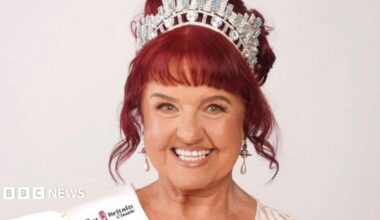 Ann Marie Harper wearing a tiara smiles at the camera. She has red hair in an updo and is wearing a white ballgown.