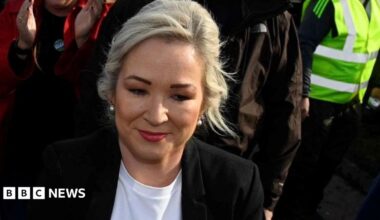 Michelle O'Neill with blonde hair, wearing a black jacket and white top. You can see part of a woman with a red coat clapping behind her, someone in a black coat and someone with a yellow hi-vis jacket to her right