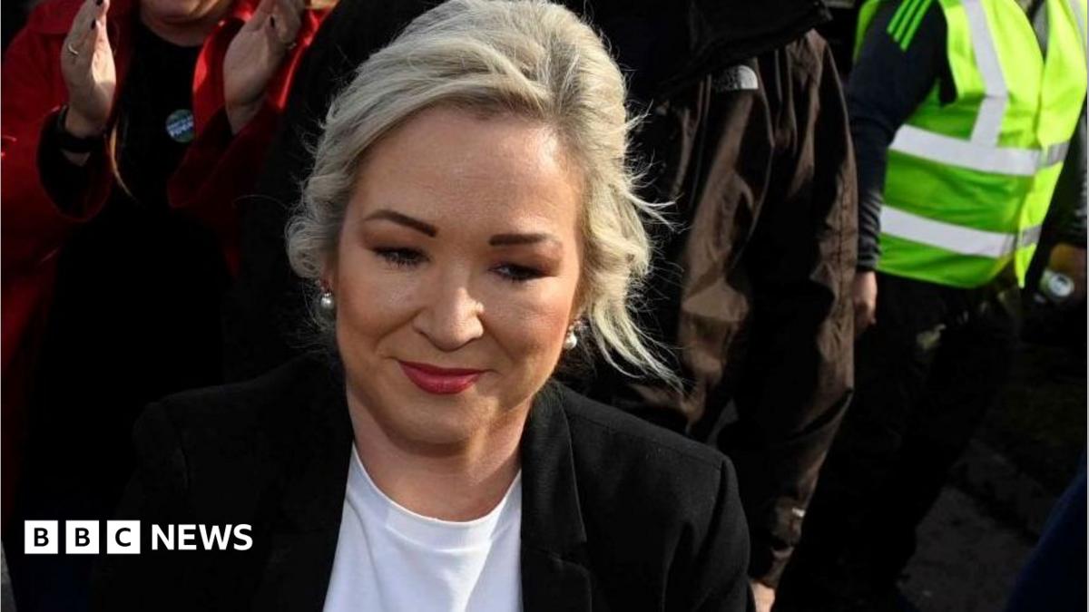 Michelle O'Neill with blonde hair, wearing a black jacket and white top. You can see part of a woman with a red coat clapping behind her, someone in a black coat and someone with a yellow hi-vis jacket to her right
