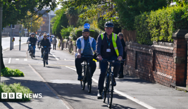 Cyclists with helmets travel along the new cycle lane. A few pedestrians are walking on the pavement next to it. The carriageway of Abbey Road is next to the cycle lane. The area is urban but not crowded. In the distance there are larger red brick buildings on the left. In the foreground, on the right, ornate and well maintained brick walls stand in front of manicured hedgerows.
