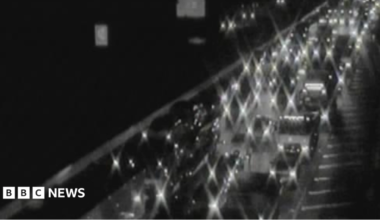 Lights of cars and vans light up in the dark with bumper to bumper traffic on one side of the M6. It is blurry black and white footage.