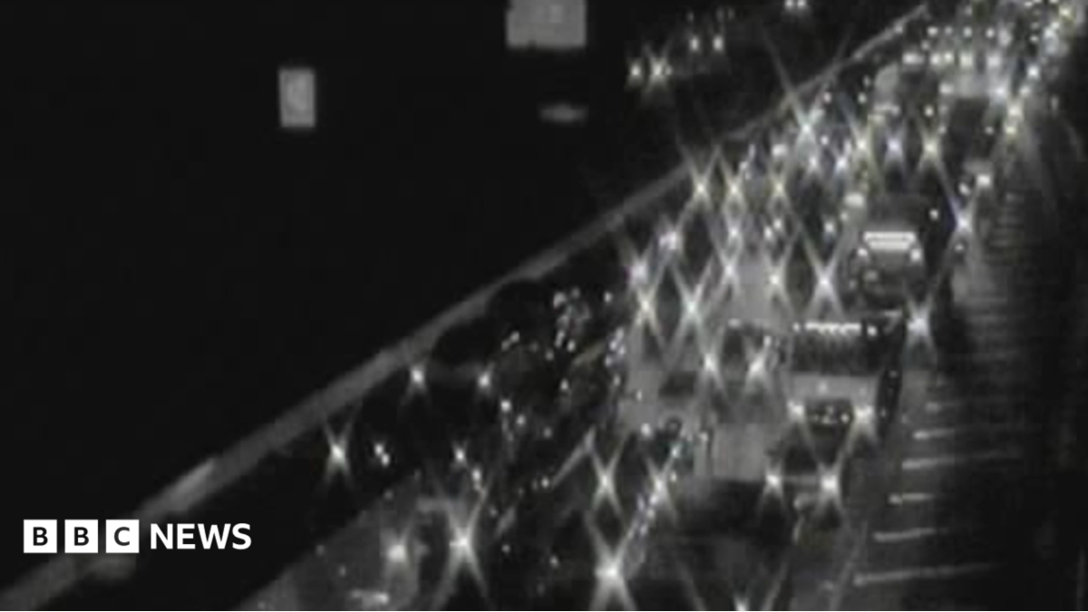 Lights of cars and vans light up in the dark with bumper to bumper traffic on one side of the M6. It is blurry black and white footage.