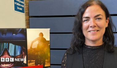 Jane Whild is outdoors in front of a red-brick building with large windows. She is wearing a dark zip-up jacket and a clip-on microphone attached to a dark top. Ornamental grasses and shrubs are visible beside a walkway in the background. A sign on the building reads “Christodoulou Meeting Rooms.”