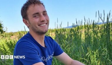 Hugo Flint Cahan is looking into the camera smiling. He is sat in a field surrounded by long grass on a bright summer day. He is wearing a blue t-shirt.