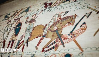 A close-up section of the Bayeux Tapestry shows embroidered figures, horses, shields and weapons in a battle scene, with decorative borders and Latin text stitched into the fabric.