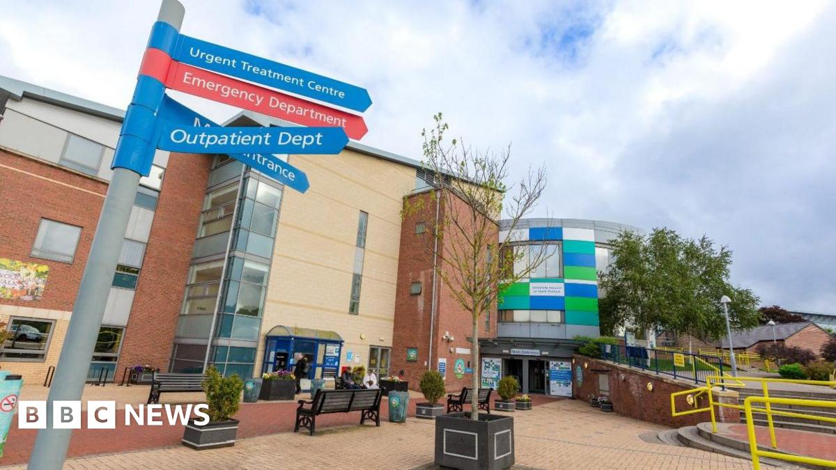 The outside of the University of North Durham Hospital. It a brown and beige-coloured brick building. The entrance has green, white and blue rectangle panelling. There is a sign pointing patients to various departments.  A small tree  and some small shrubs are in plant pots in a seating area. There are yellow railings around some steps to the right.