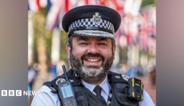 A man in a Metropolitan Police uniform and peaked cap stands outdoors, wearing a radio and body-worn camera on a protective vest. Red, white and blue flags are visible blurred in the background.
