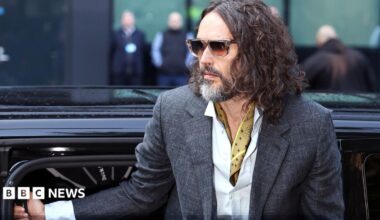 Russell Brand gets out of a car wearing brown sunglasses, a grey suit with a yellow fabric around as his neck. He has long hair down to his shoulders.