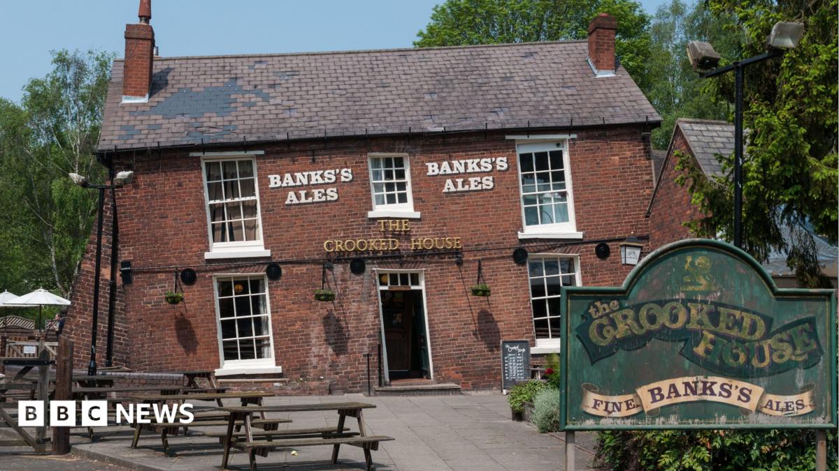 Th crooked house pub - a slanted red brick pub with a sign outside and picnic benches