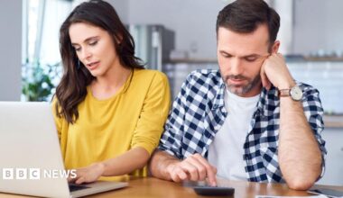 Couple sit at a kitchen table with the woman in a yellow jumper looking at a laptop and the man in a blue and white shirt pressing numbers on a calculator.