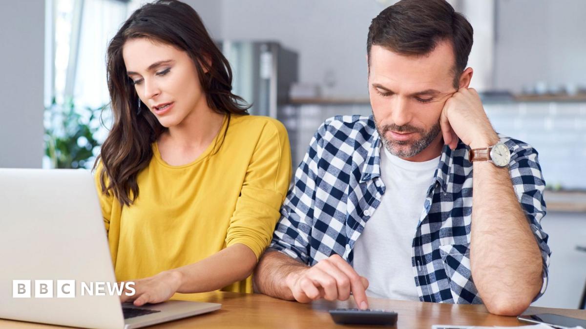 Couple sit at a kitchen table with the woman in a yellow jumper looking at a laptop and the man in a blue and white shirt pressing numbers on a calculator.