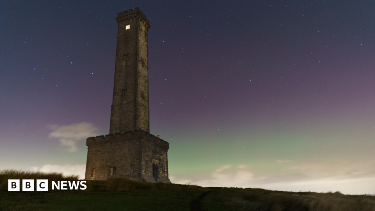 The Peel Tower stands on a hillside beneath a star-filled night sky, illuminated by a green and purple aurora glow on the horizon. Low clouds drift across the landscape below the historic stone monument.