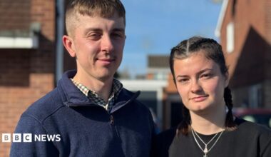 Ben and Meg stand outside. Ben is wearing a navy quarter zip top and checked shirt. Meg is wearing a black top and necklace.