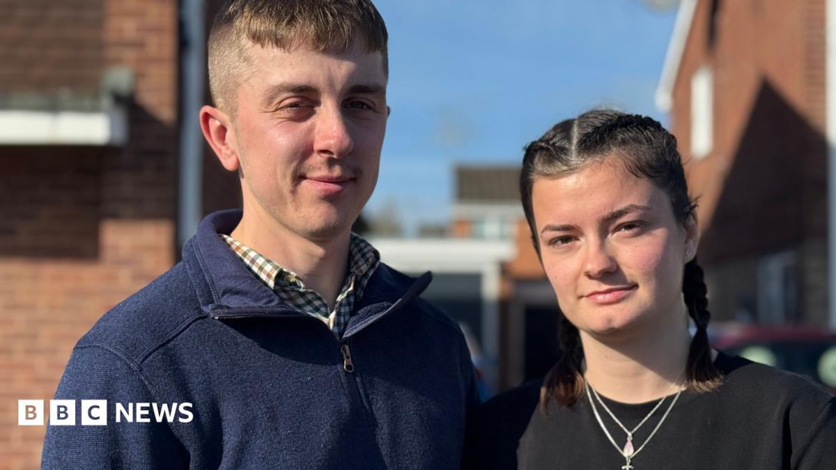 Ben and Meg stand outside. Ben is wearing a navy quarter zip top and checked shirt. Meg is wearing a black top and necklace.