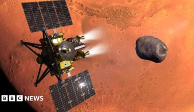 An artist's impression of the MMX spacecraft - which is a gold and black spacecraft with large solar panels, thrusters and antenna - flies above the red surface of Mars, which looms large in the background. It is heading towards the small, dark and irregularly shaped moon called Phobos.