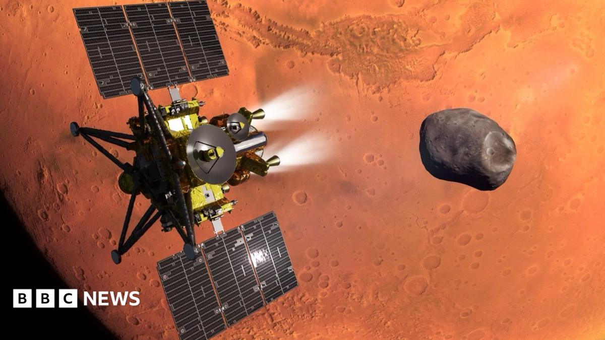 An artist's impression of the MMX spacecraft - which is a gold and black spacecraft with large solar panels, thrusters and antenna - flies above the red surface of Mars, which looms large in the background. It is heading towards the small, dark and irregularly shaped moon called Phobos.