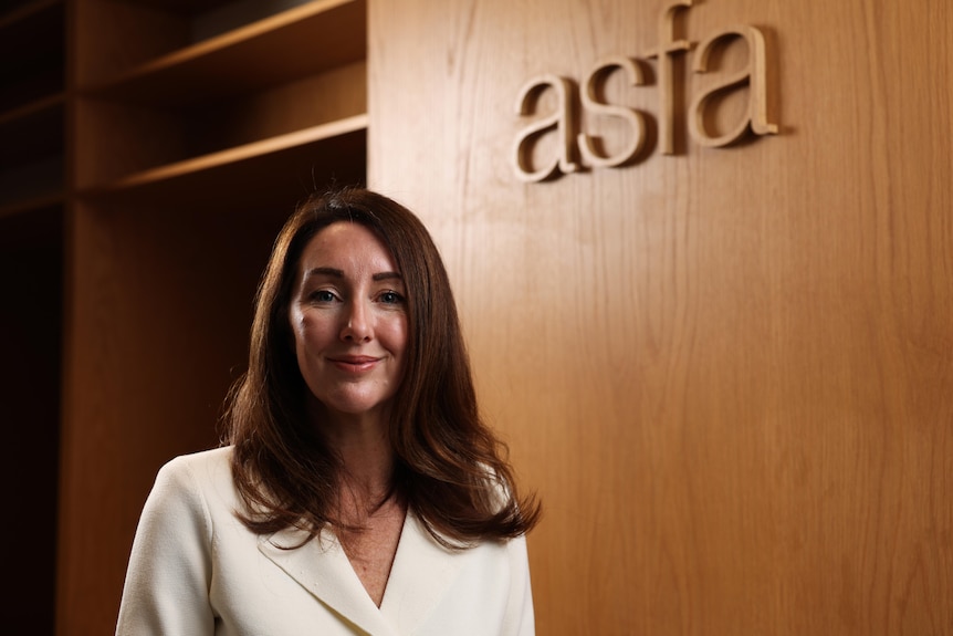 Mary Delahunty wears a brown dress-jacket and stands in front of a wooden wall with a plaque that reads ASFA.