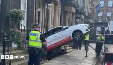 A car being lifted out from the basement level of a flat building by a tow truck, which is off to the right side of the picture on the road. The car is white with red writing on the side and a red line at the bottom. Three Police Scotland officers stand and watch nearby