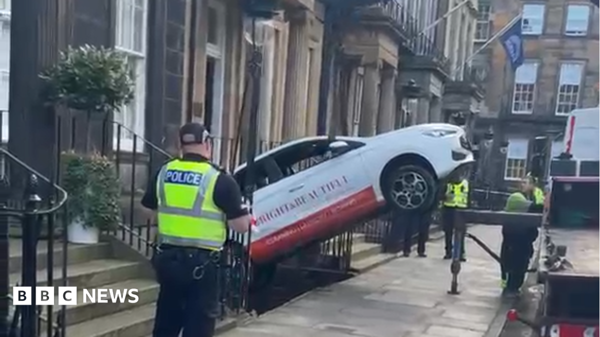 A car being lifted out from the basement level of a flat building by a tow truck, which is off to the right side of the picture on the road. The car is white with red writing on the side and a red line at the bottom. Three Police Scotland officers stand and watch nearby