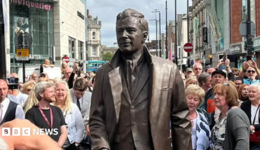 A bronze statue of Brian Epstein wearing a suit man in the centre of a busy Liverpool city street, surrounded by a large crowd of spectators and photographers during an unveiling event. Shops and traffic signs are visible in the background under a bright daytime sky.