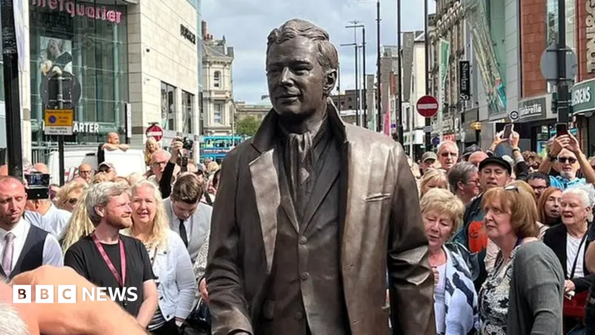 A bronze statue of Brian Epstein wearing a suit man in the centre of a busy Liverpool city street, surrounded by a large crowd of spectators and photographers during an unveiling event. Shops and traffic signs are visible in the background under a bright daytime sky.