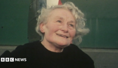 Margaret smiling, staring just above the camera. She is stood in front of a Spitfire. She has short, curly, grey hair and is wearing a black sweater.