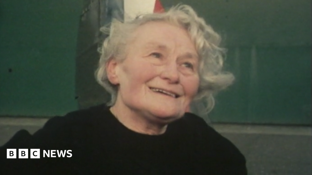 Margaret smiling, staring just above the camera. She is stood in front of a Spitfire. She has short, curly, grey hair and is wearing a black sweater.