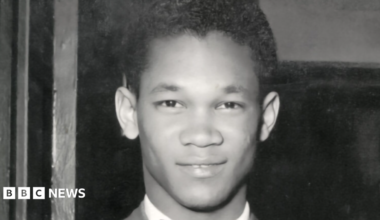 Black-and-white portrait photograph of Lord Woodbine wearing a suit jacket and a patterned bow tie. The image is a close-up head-and-shoulders shot taken indoors, with dark wooden panels visible in the background.